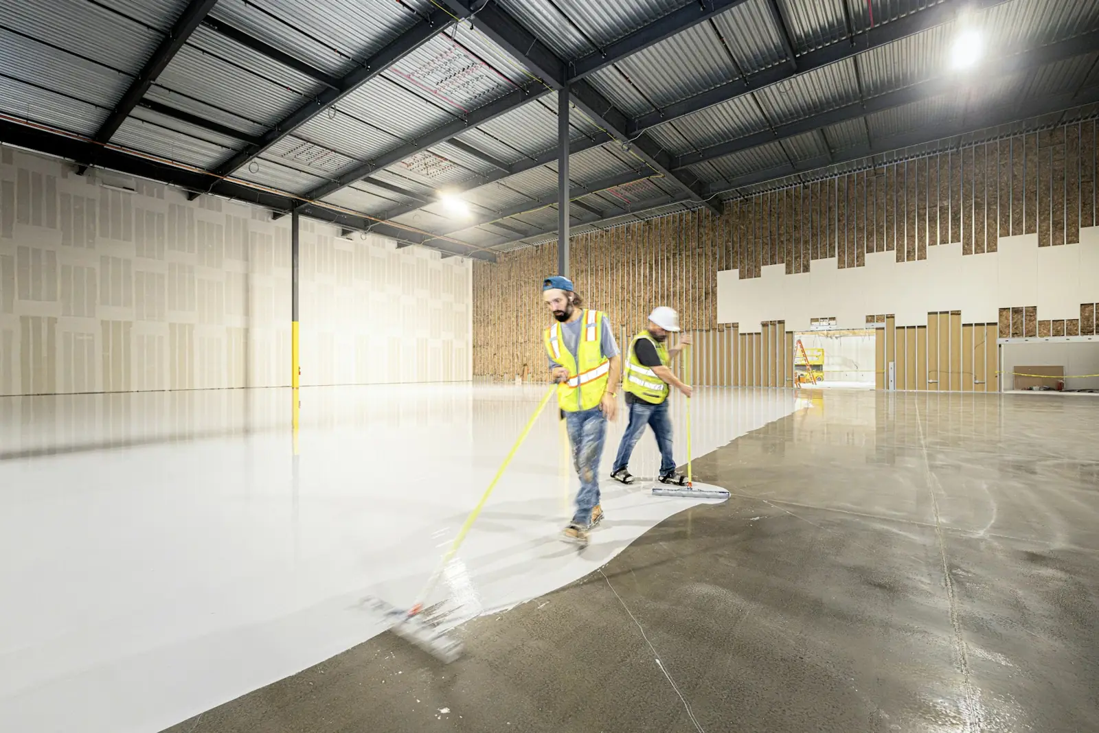 Two installers spreading white epoxy coating across a large concrete floor with squeegees