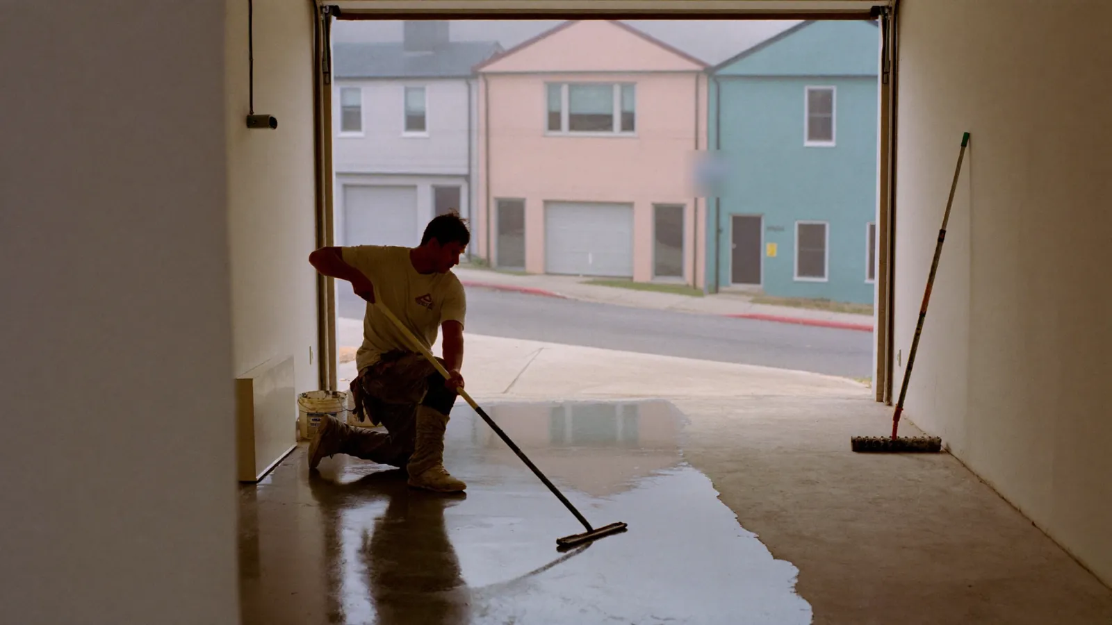Installer kneeling to squeegee gray epoxy across a narrow garage floor with the roll-up door open to the street
