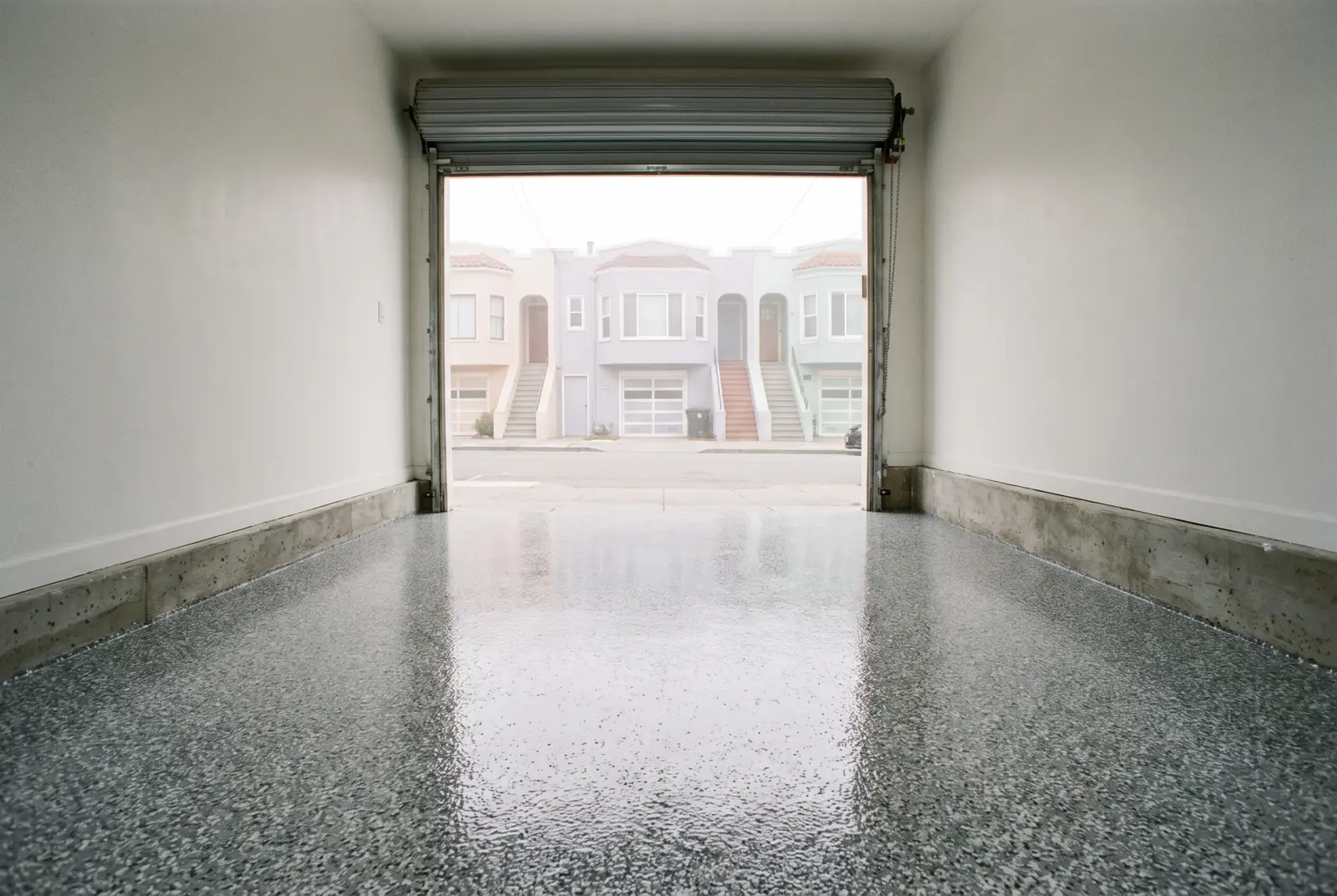 Glossy gray flake epoxy floor in a narrow garage with the door open to a row of houses